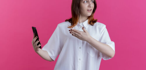 looking at side unpleased young redhead girl holding and points with hand at phone isolated on pink background